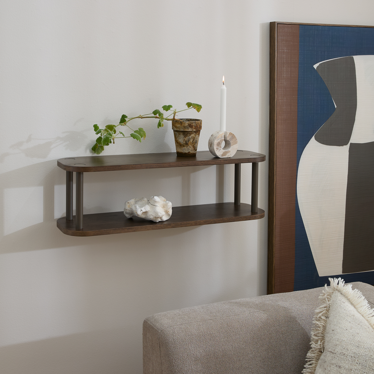 Væghylde i mørkt træ med to hylder; på hylden står en grøn plante i potte, et rustikt kar, et hvidt stearinlys og en lille stenskulptur, ved siden af et abstrakt kunstværk og en beige sofa.