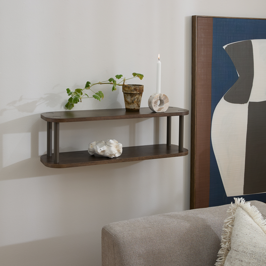Væghylde i mørkt træ med to hylder; på hylden står en grøn plante i potte, et rustikt kar, et hvidt stearinlys og en lille stenskulptur, ved siden af et abstrakt kunstværk og en beige sofa.