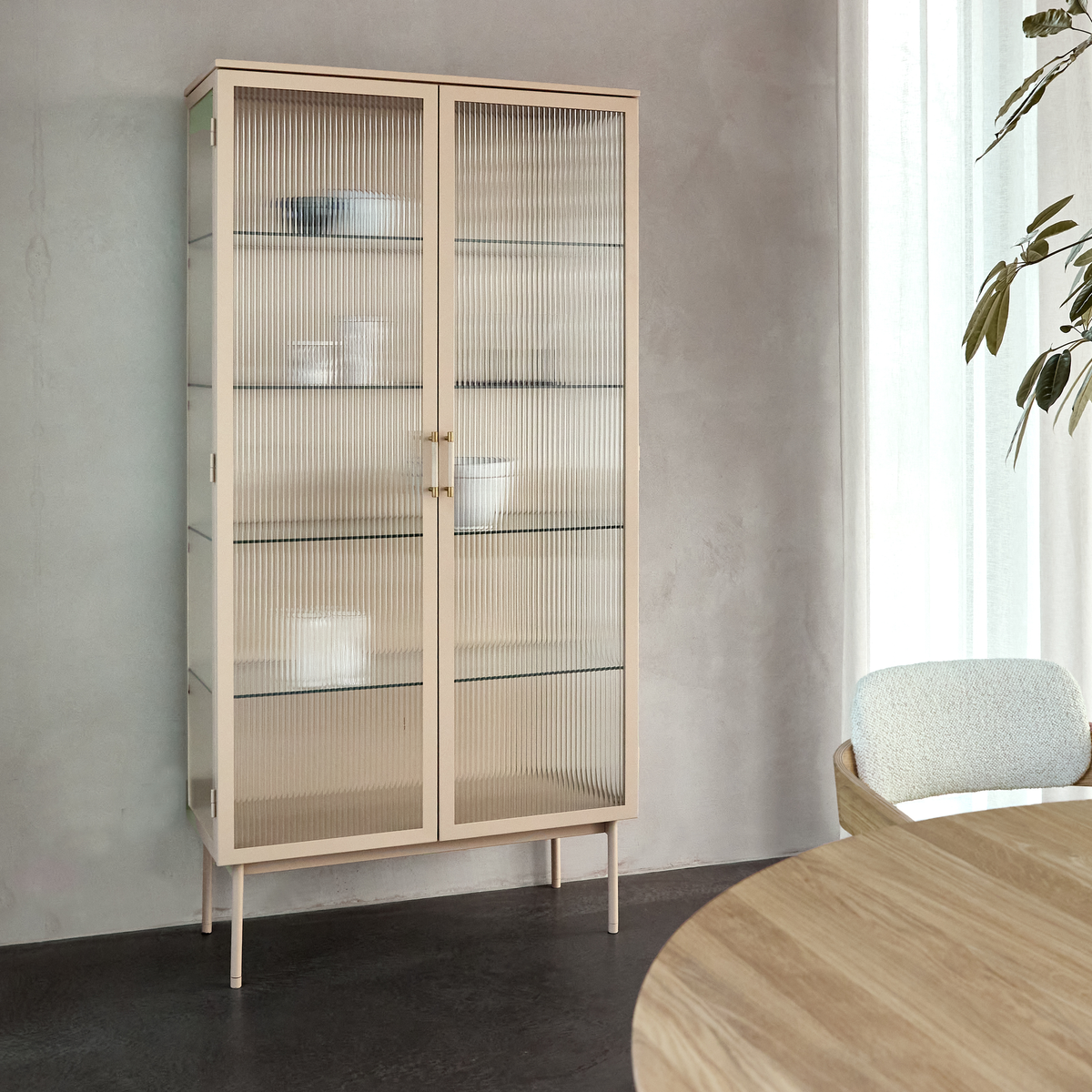 Beige vitrineskab med rillet glas og slanke ben i et lyst spisemiljø; rund træbordplade og let gardin i dagslys.
