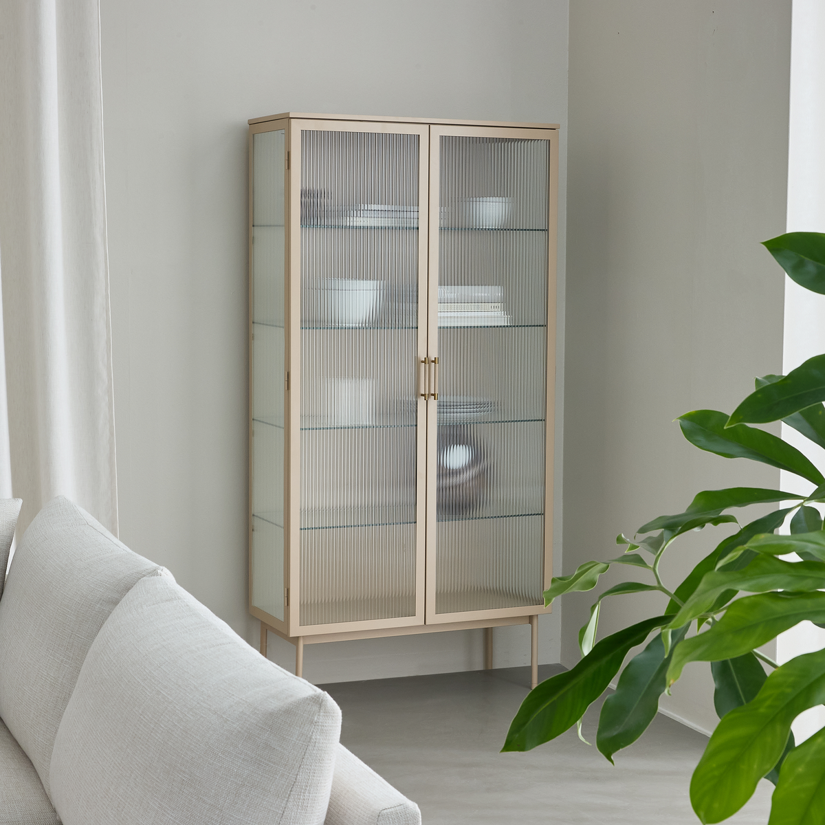 Lyst vitrineskab med riflede glaslåger i en minimalistisk stue, med hvide skåle og bøger på glashylder og en grøn plante ved siden af.