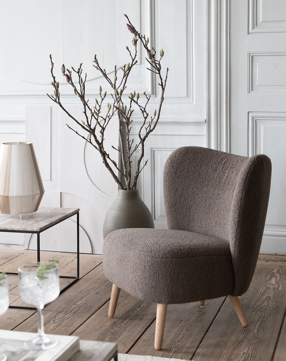 Hyggelig loungestol i brun bouclé med træben ved siden af stor vase med nøgne grene, lille bordlampe og marmorbord på trægulv i lyst stuemiljø.