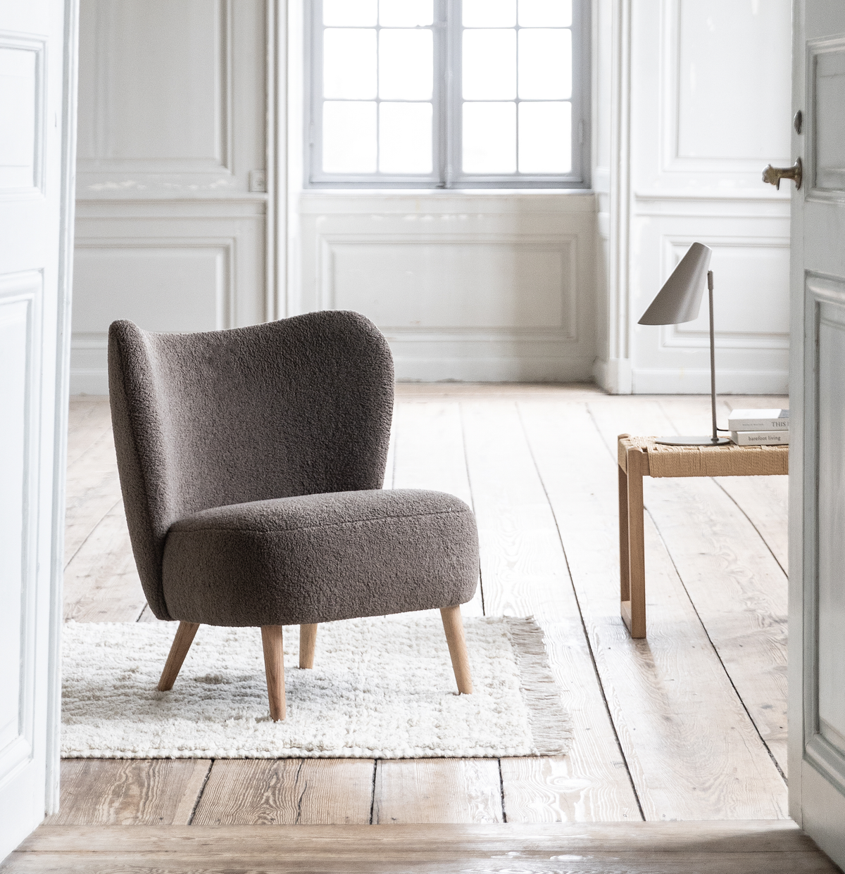 Lyst interiør med en grå bouclé-loungestol på et hvidt ryetæppe, trægulv og et lille træbord med en minimalistisk bordlampe ved siden af et vindue.
