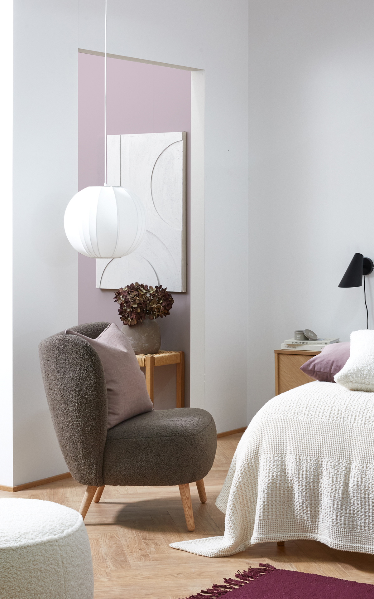 Lyst soveværelse i skandinavisk stil med grå lænestol, hvid pendellampe, seng med hvid strikket plaid og lilla accenter samt tørrede blomster på et lille bord.