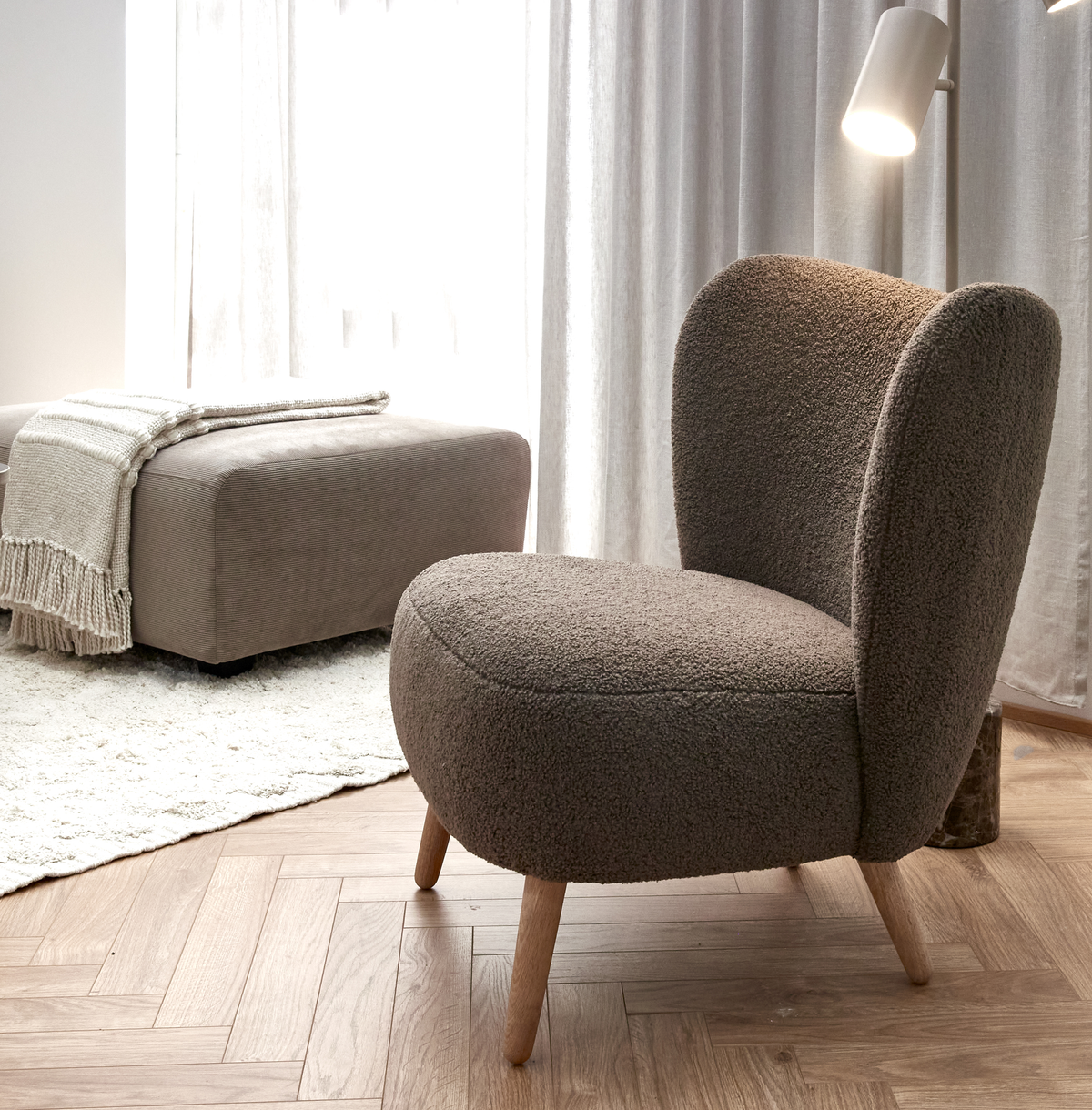 Beige-brun loungestol i bouclé med træben i en lys stue, med gulvlampe, puf med plaid og lyse gardiner.