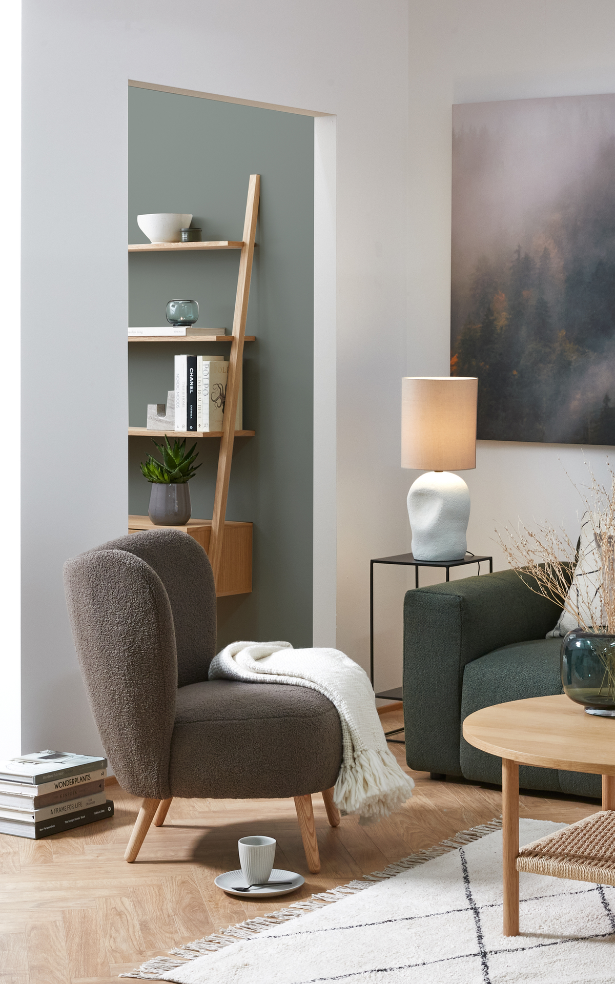 Moderne skandinavisk stue med grøn sofa, brun lænestol med hvid plaid, rundt sofabord i træ, tænd bordlampe og bogstakke med titlerne CHANEL, WONDERPLANTS og A FRAME FOR LIFE i rolige, neutrale toner.