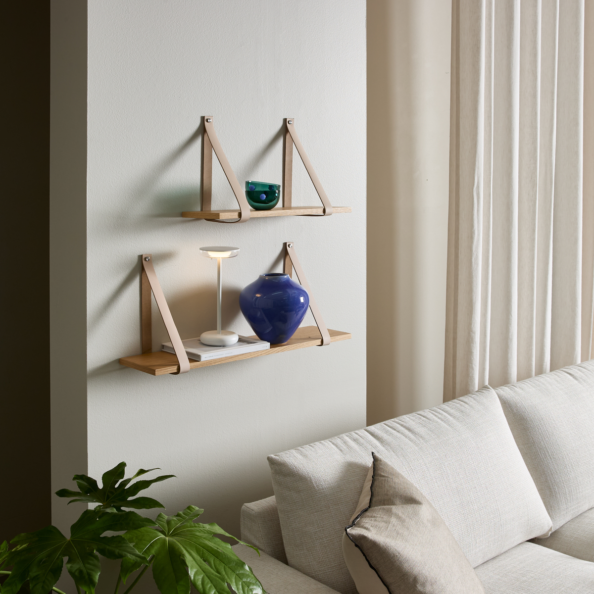 Væghylder med læderstropper over en lys sofa; lille bordlampe, blå vase og bøger, beige gardiner og grøn plante.