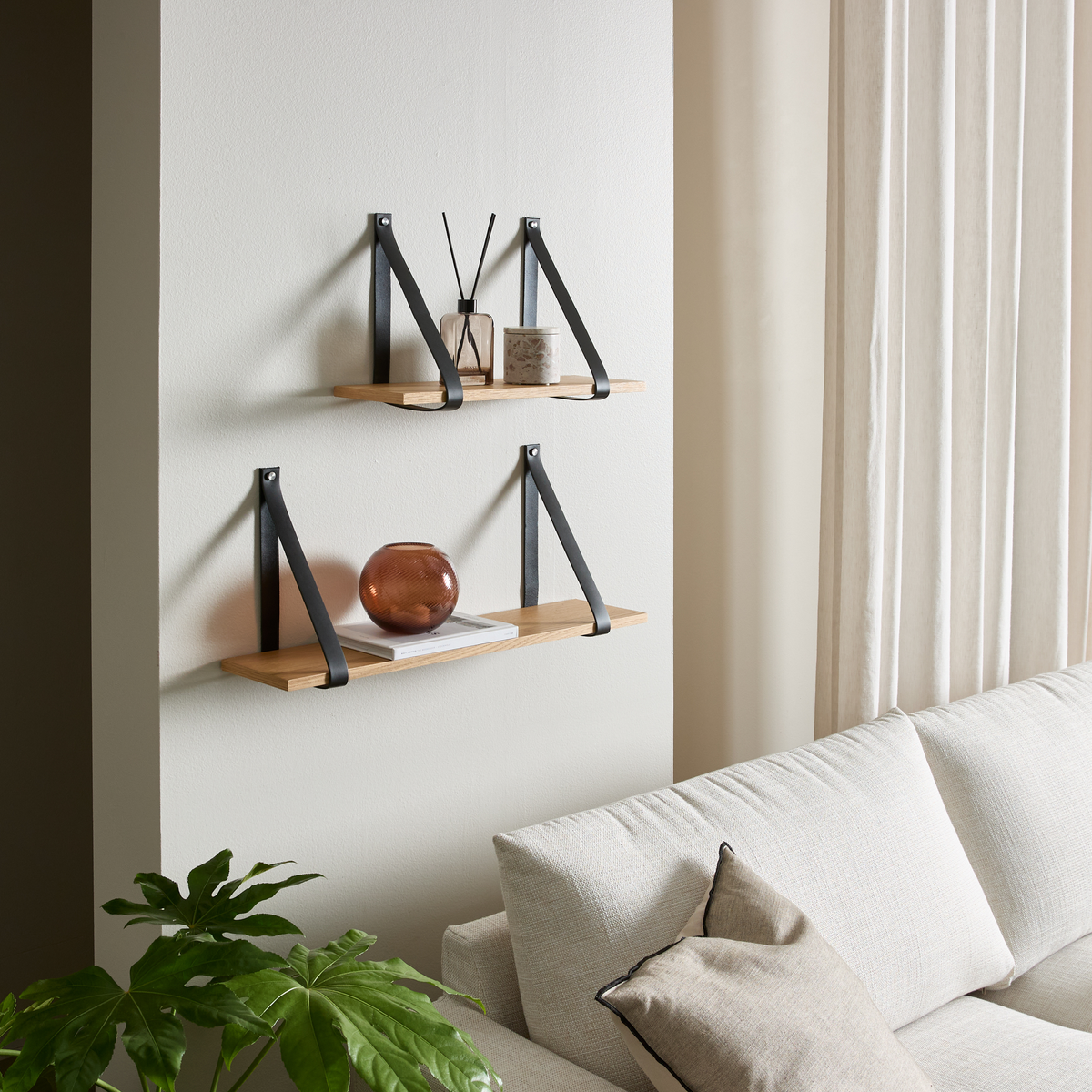 Vægmonterede træhylder med sorte beslag over en lys sofa, stylet med diffuser, lys, vase og bøger, med lyse gardiner og grøn plante i en lys stue.
