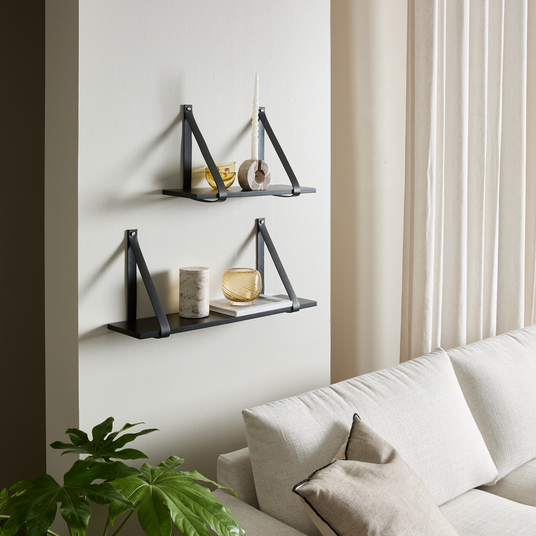 To sorte væghylder med minimalistisk pynt – snoet lys, stenring, glasvase og skål – over en lys sofa ved siden af lyse gardiner og en grøn plante.