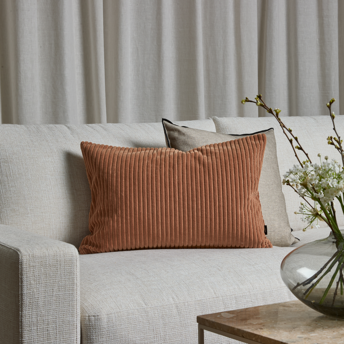 Beige sofa med rillet terrakottafarvet rektangulær pude og en neutral pude bagved foran lyse gardiner; glasvase med grene på stentopbord.