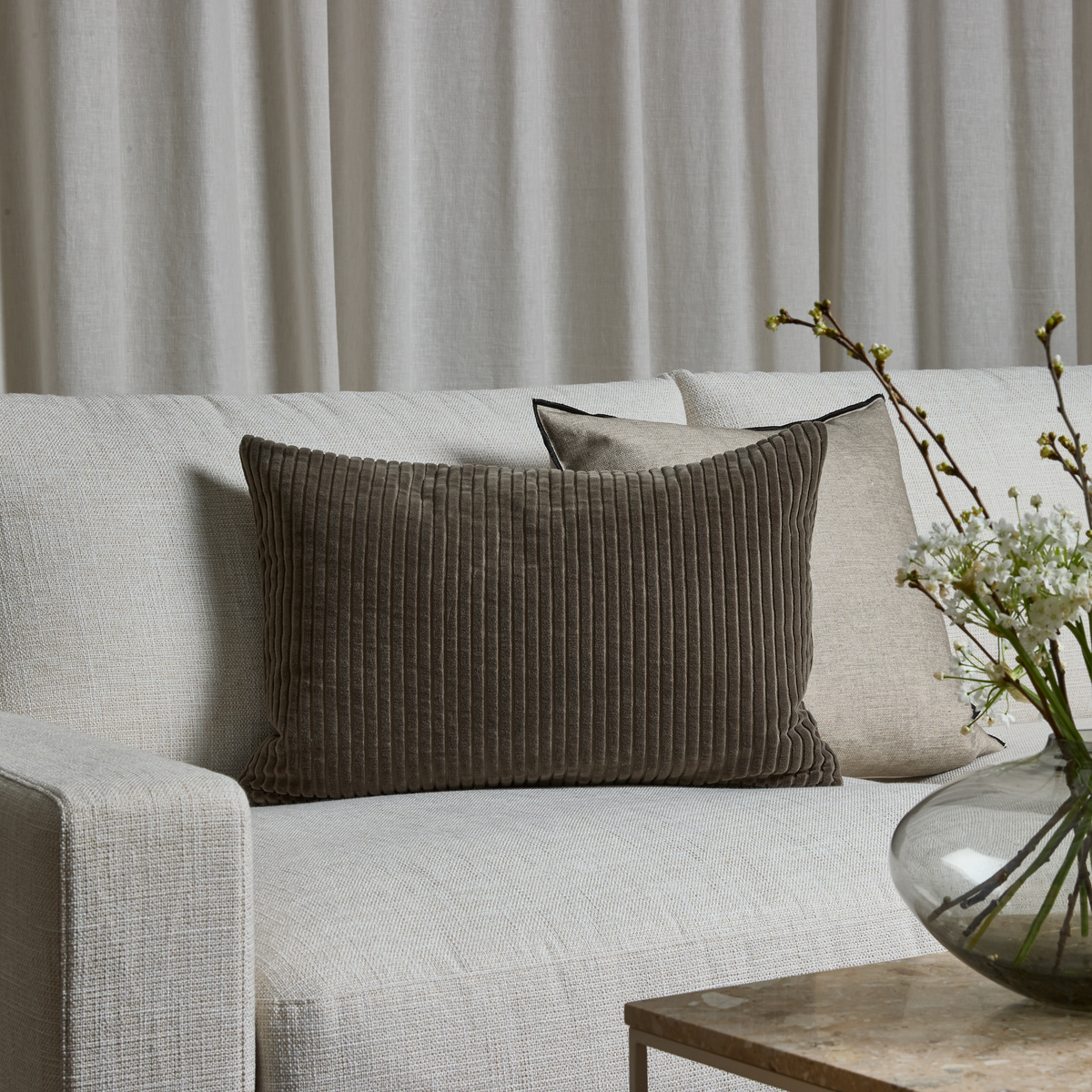 Lys beige sofa med en mørkebrun rillet, rektangulær pude og to neutrale puder, lyse gardiner bagved samt glasvase med hvide blomster på et sidebord.