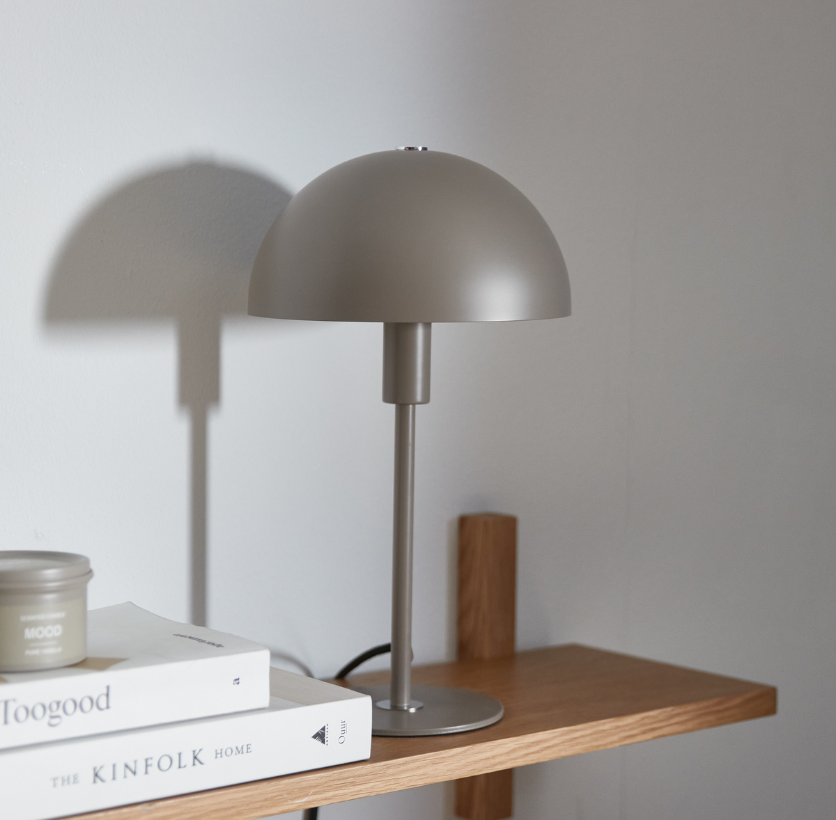 Beige kuppelformet bordlampe på en træhylde ved siden af et lille lys med teksten MOOD og to bøger, Toogood og The Kinfolk Home.