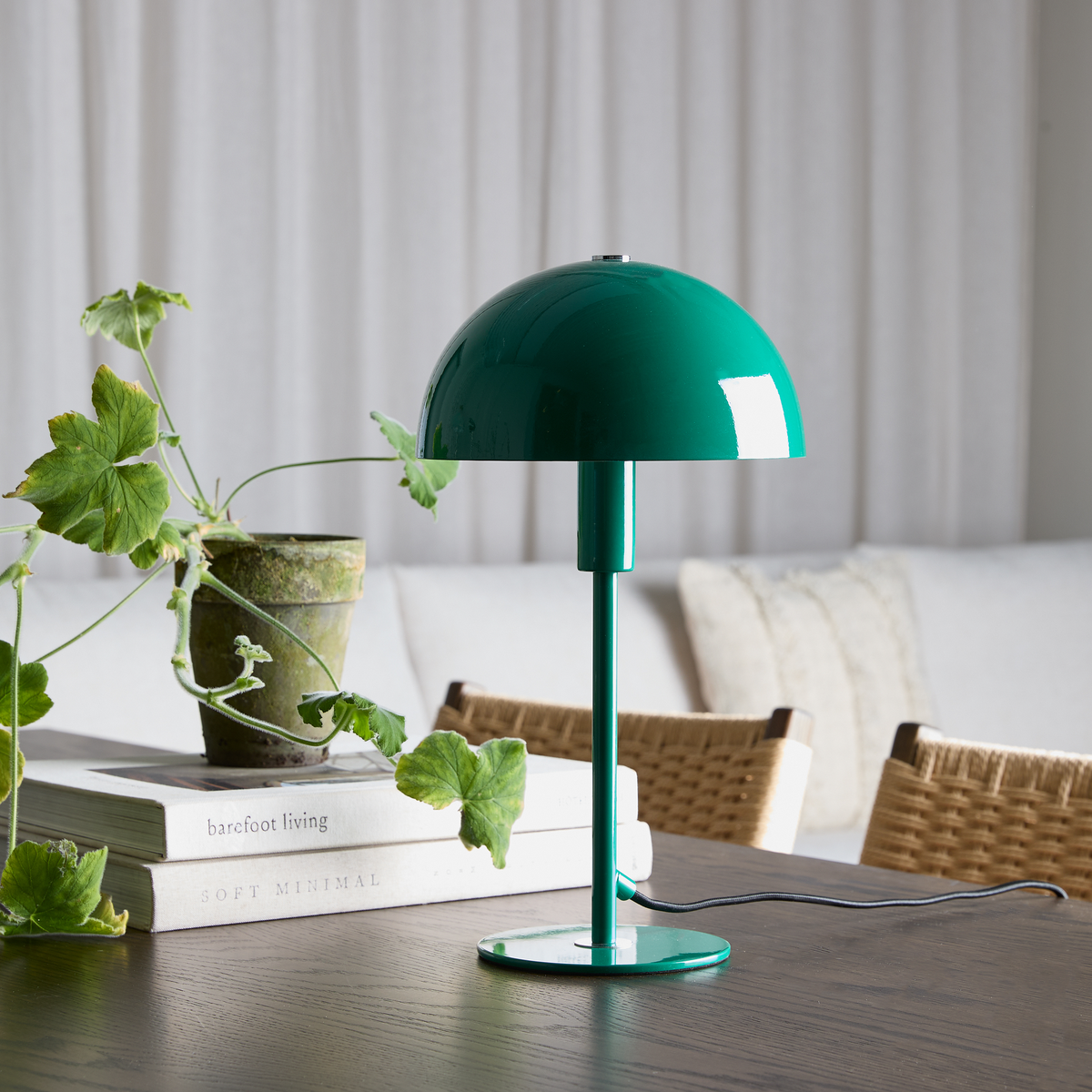 Grøn bordlampe med halvkuplet skærm på mørkt bord ved siden af en potteplante og bøgerne Barefoot Living og Soft Minimal i en lys skandinavisk stue.