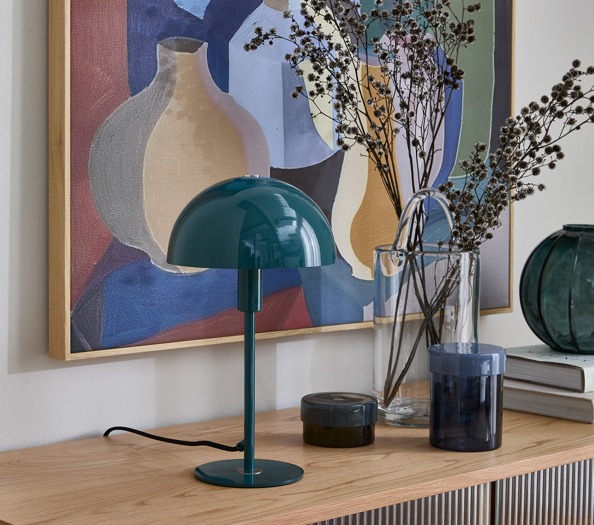 Turkisgrøn kuppelformet bordlampe på træskænk med glasvaser, bøger og abstrakt vægkunst.