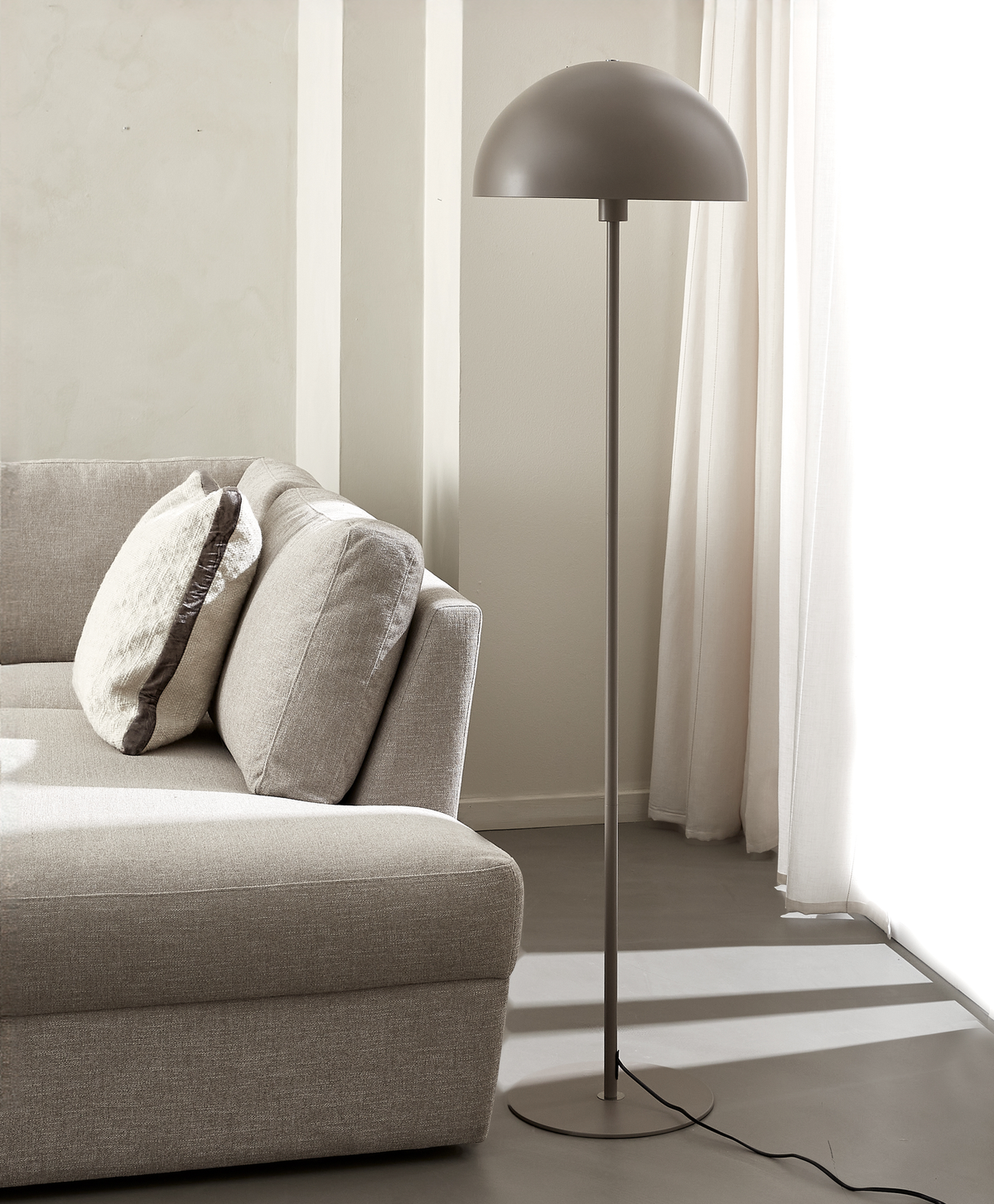 Moderne gulvlampe med kuppelformet skærm ved siden af beige sofa i lys stue med hvide gardiner.