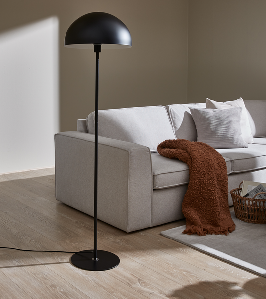 Sort gulvlampe med halvkupet skærm står ved siden af en lysegrå sofa med puder, en brun plaid og en flettet kurv på et lyst tæppe i blødt dagslys.