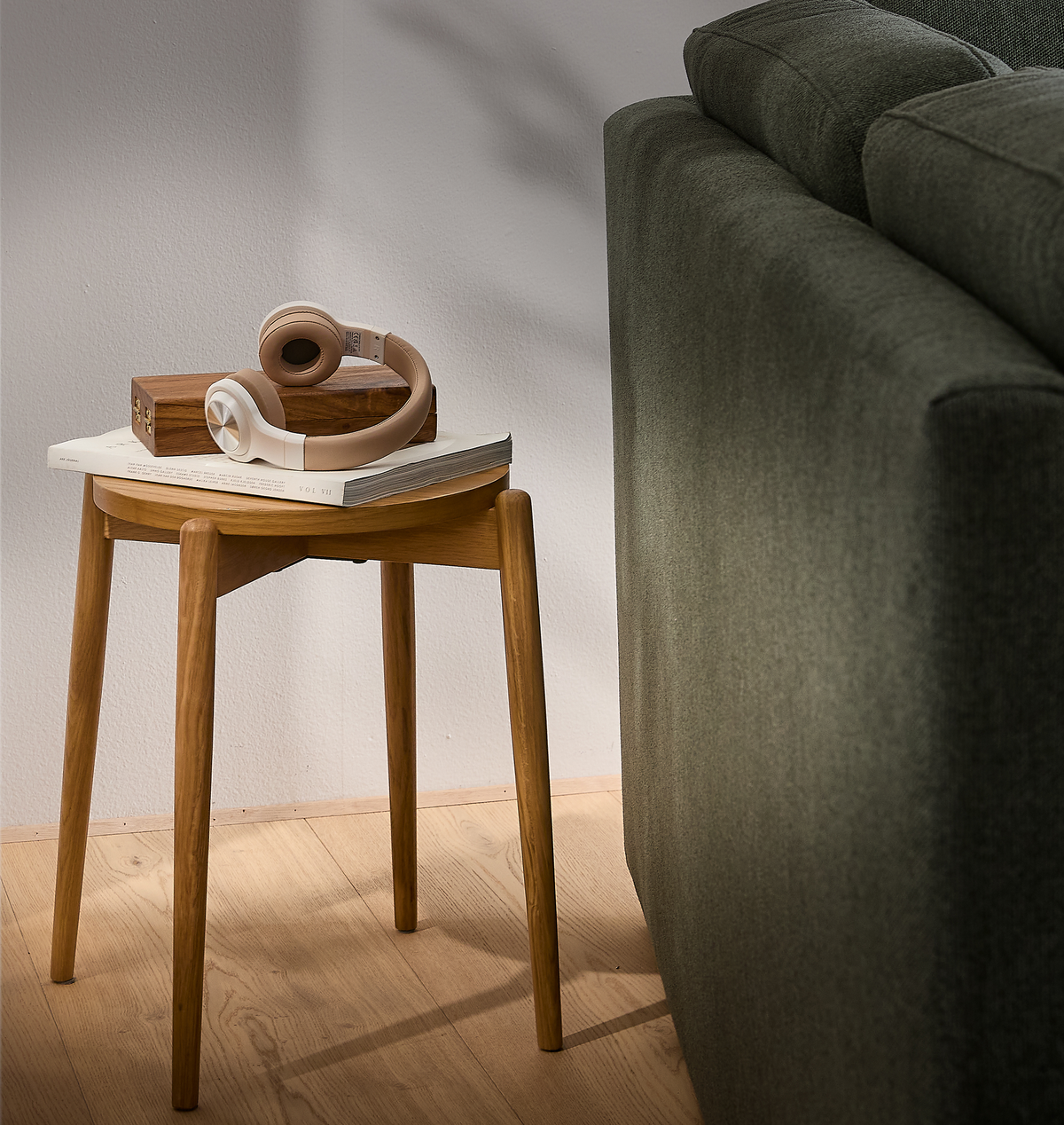 Lille rundt sidebord i lyst egetræ ved siden af en grøn sofa, med beige hovedtelefoner, en trææske og en bog med titlen VOL VII i blødt dagslys.
