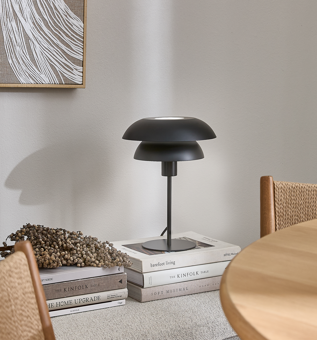 Sort bordlampe på en stak bøger med titlerne Barefoot Living, The Kinfolk Table, The Home Upgrade og Soft Minimal, ved siden af en træstol og et rundt spisebord i en lys skandinavisk indretning.