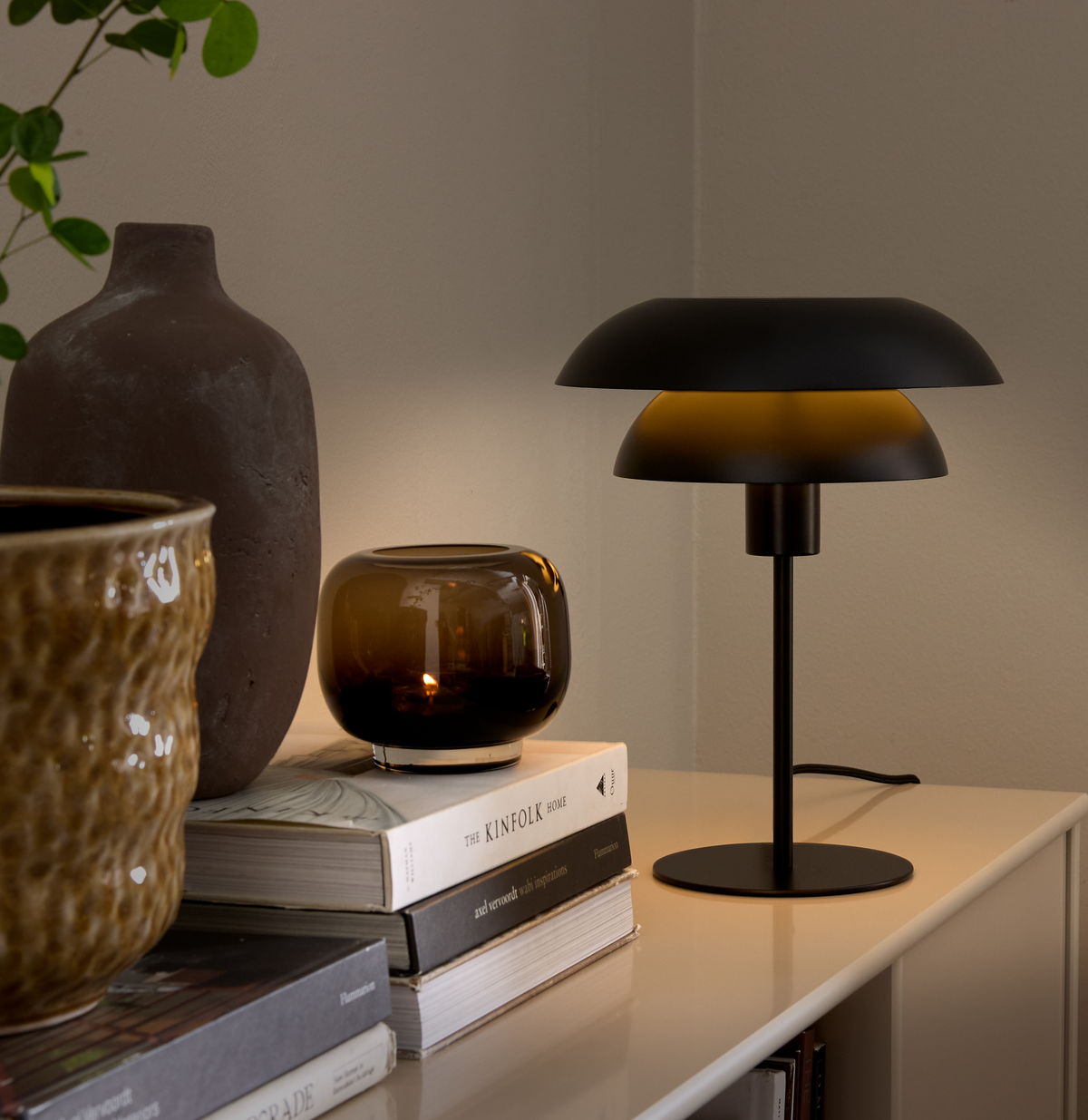Sort bordlampe med varm glød ved siden af en ravfarvet lyslygte på en stak bøger, bl.a. The Kinfolk Home, på en skænk med keramiske vaser.