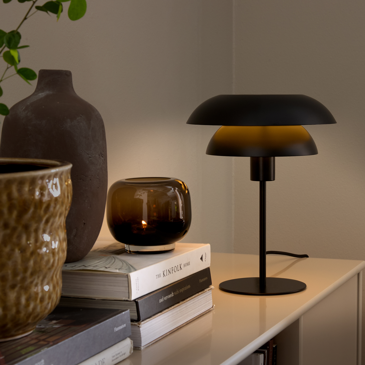 Sort bordlampe på en skænk ved siden af en ravfarvet glasstage med tændt flamme, keramikvaser og en stak bøger med titlen The Kinfolk Home.