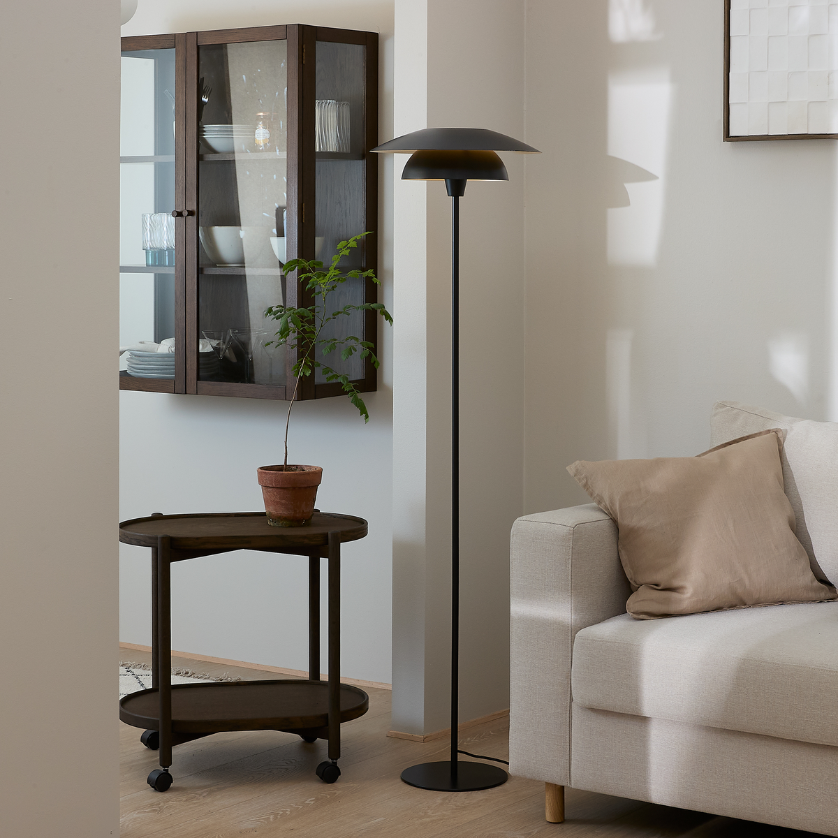Lyst stue med beige sofa, sort gulvlampe, rullebord i træ med potteplante og væghængt vitrineskab.