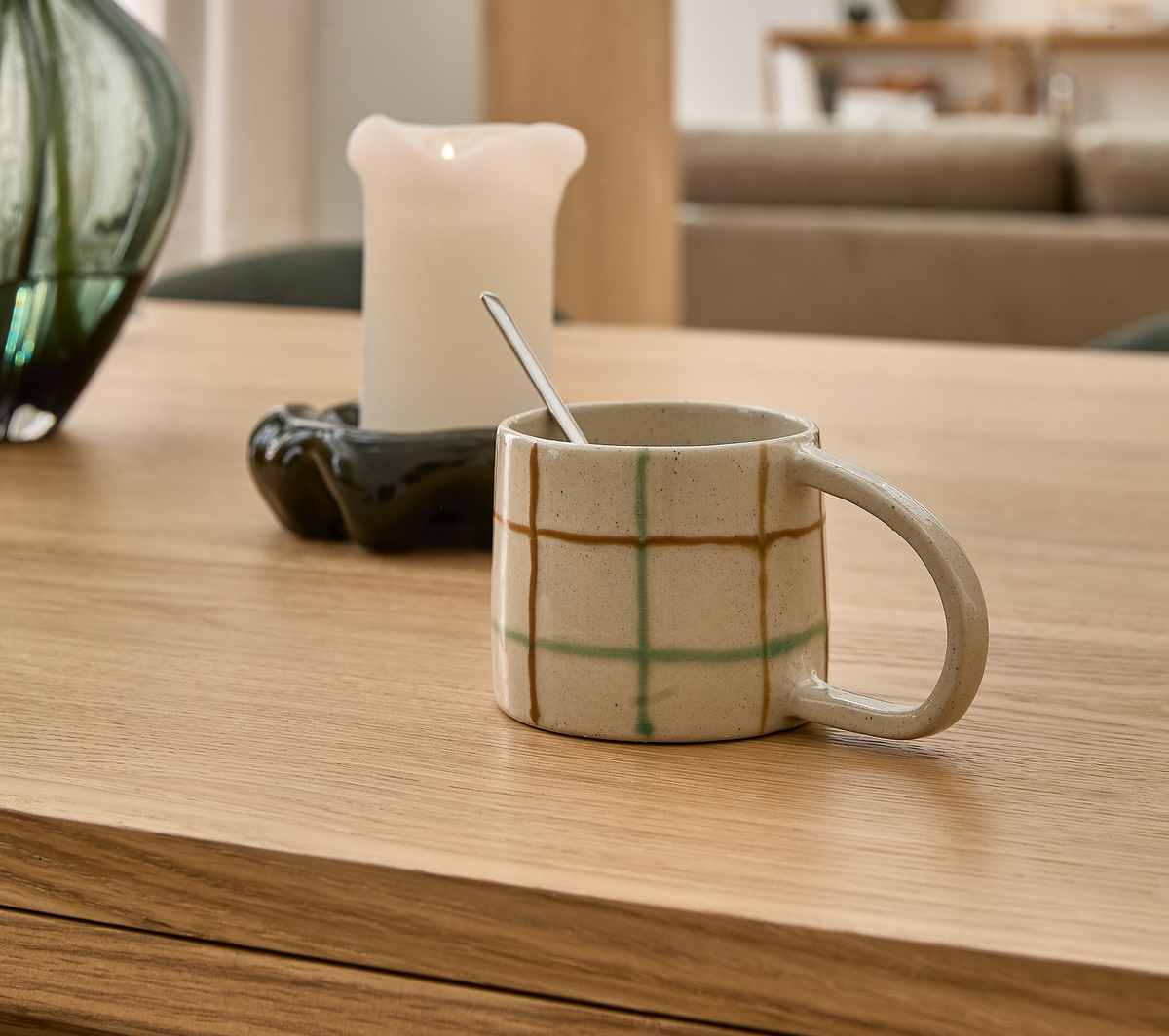 Keramikkop med ternet mønster og ske på et lyst træbord, med tændt lys og grøn vase i en rolig stue.