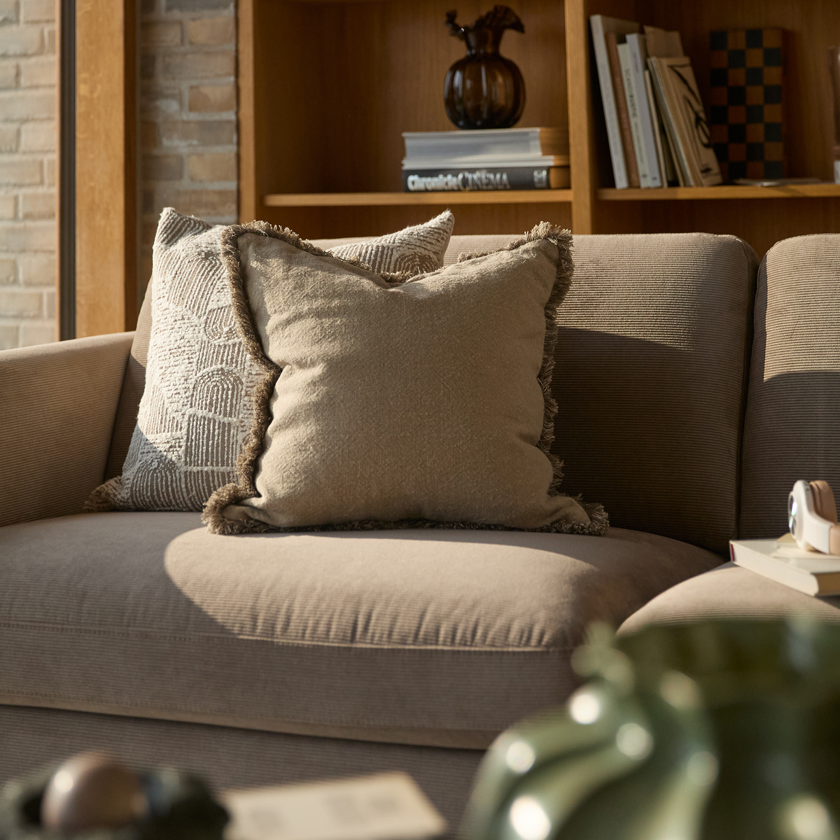 Beige sofa med to puder i sollys, træreol med vase og bogen ”Chronicle of Cinema” i baggrunden.