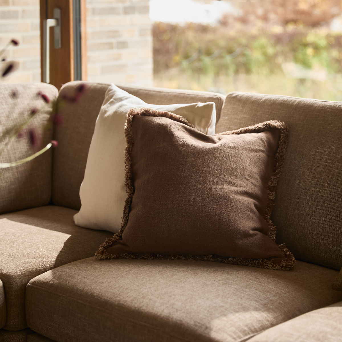 Brun pude med frynset kant på beige sofa i varmt sollys ved et vindue.
