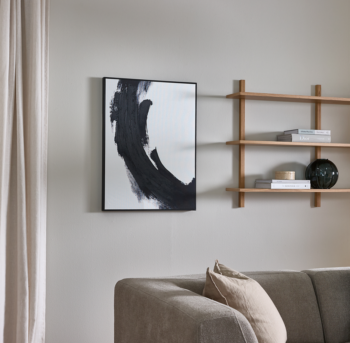 Minimalistisk stue med grå sofa, beige pude, abstrakt sort-hvid vægkunst og væghylder med bøgerne Dior og Kinfolk samt en mørk glasvase.