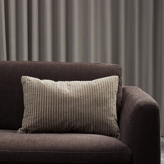 Beige rillet pude på mørkebrun sofa foran grå gardiner.