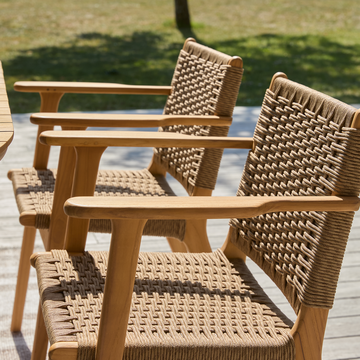 To udendørs armstole i træ med flettet sæde og ryg på en solrig terrasse.