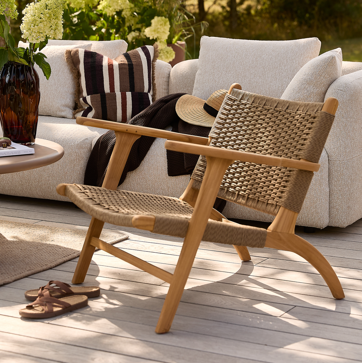 Lav loungestol i træ med flettet sæde på lys terrasse, foran beige sofa med puder, plaid og stråhat; sommerlig udendørs lounge med vase og sandaler i sollys.