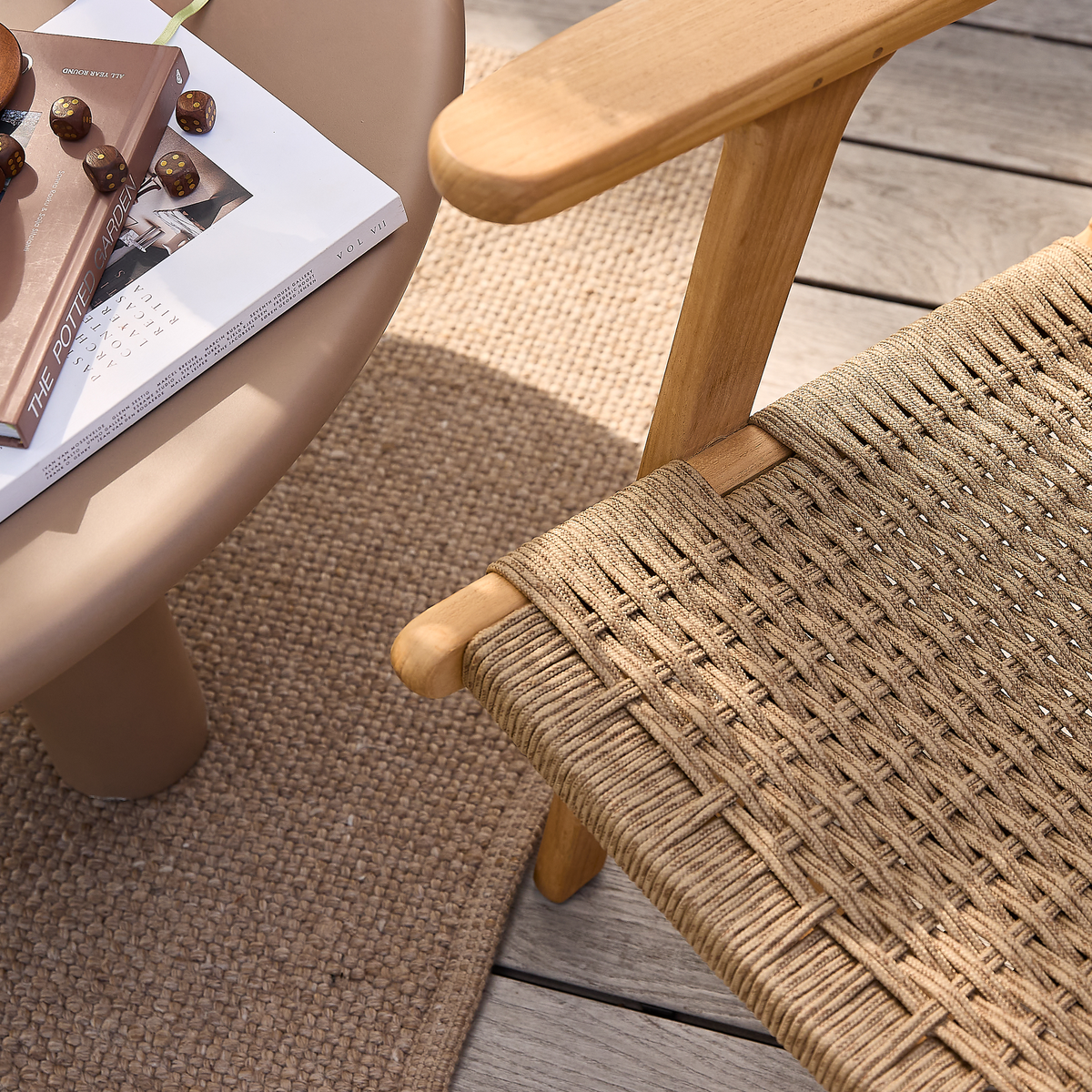 Nærbillede af flettet naturfiberstol i træ ved siden af rund beige bordplade med The Potter’s Apprentice, Vol VII og chokoladepraliner på en jutemåtte på terrasse i dagslys.