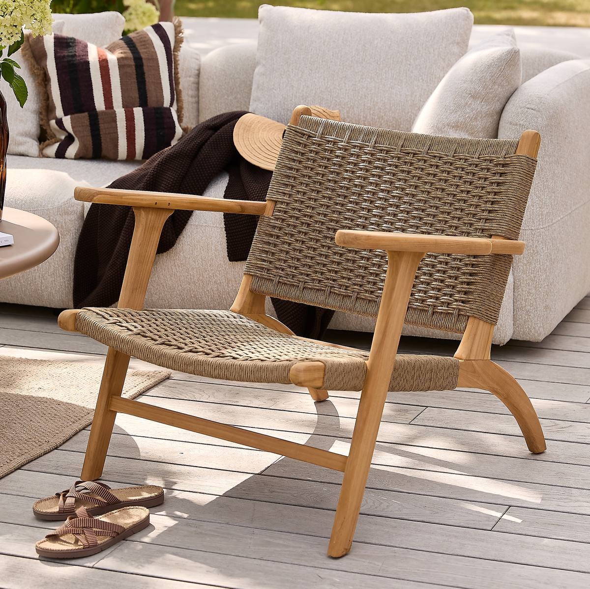 Lav loungestol i træ med flettet sæde og ryg på en solrig terrasse, foran en beige sofa med puder og plaid; sandaler ligger på trædækket.