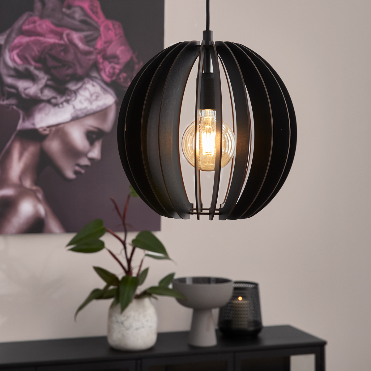 Sort kugleformet pendellampe med synlig pære lyser over et skænk med plante, skål og lys i en moderne stue.