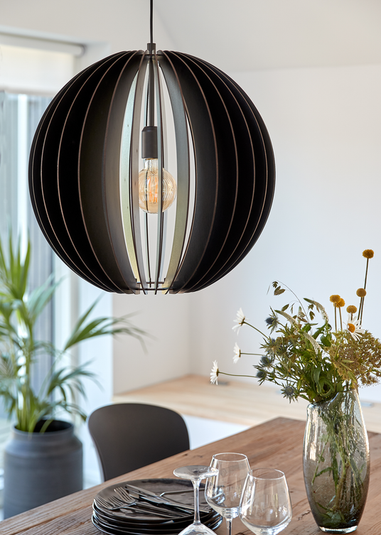 Sort rund pendel med synlig pære over et træbord med glas, sorte tallerkener og en vase med vilde blomster i en lys spisestue.