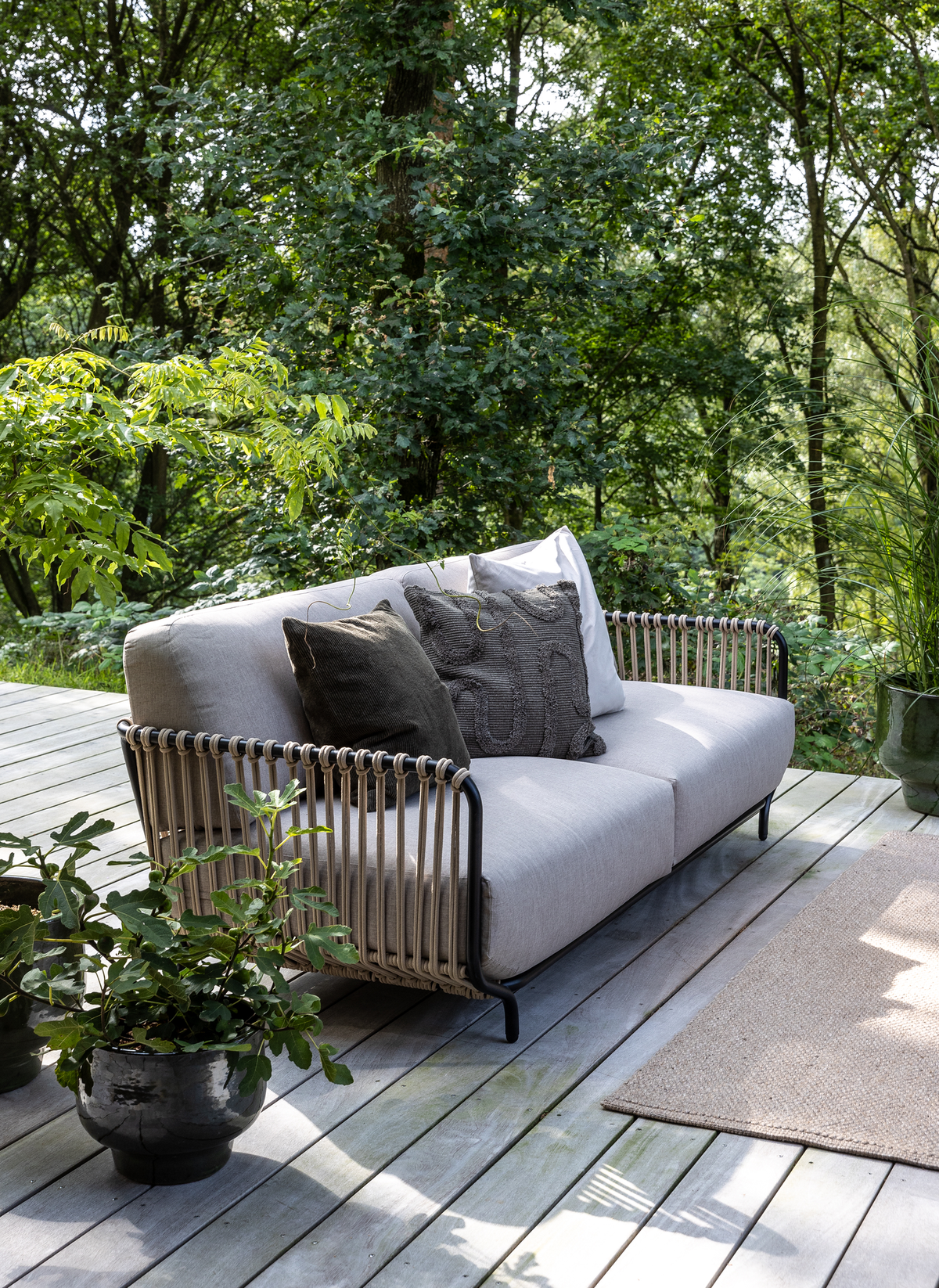 Lysegrå udendørs sofa med flettede armlæn og mørke puder på træterrasse, omgivet af grønne planter i dagslys.