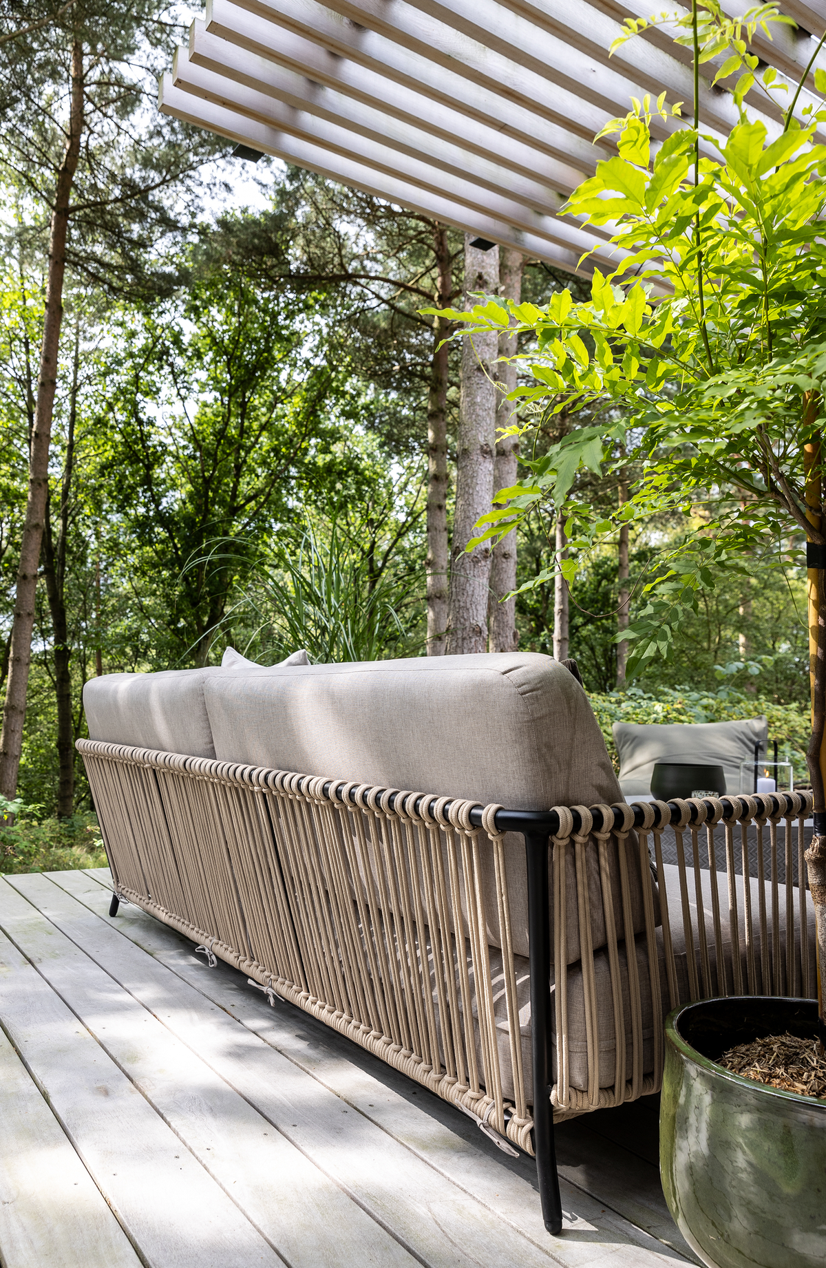 Udendørs terrasse med beige sofa med rebdetaljer under en lamel-pergola, omgivet af grønne træer; krukkeplante og lille bord med lys.