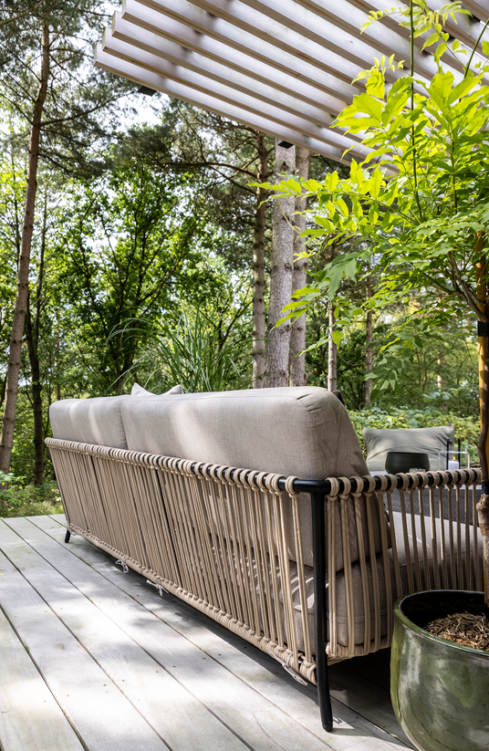 Udendørs terrasse med beige sofa med rebdetaljer under en lamel-pergola, omgivet af grønne træer; krukkeplante og lille bord med lys.