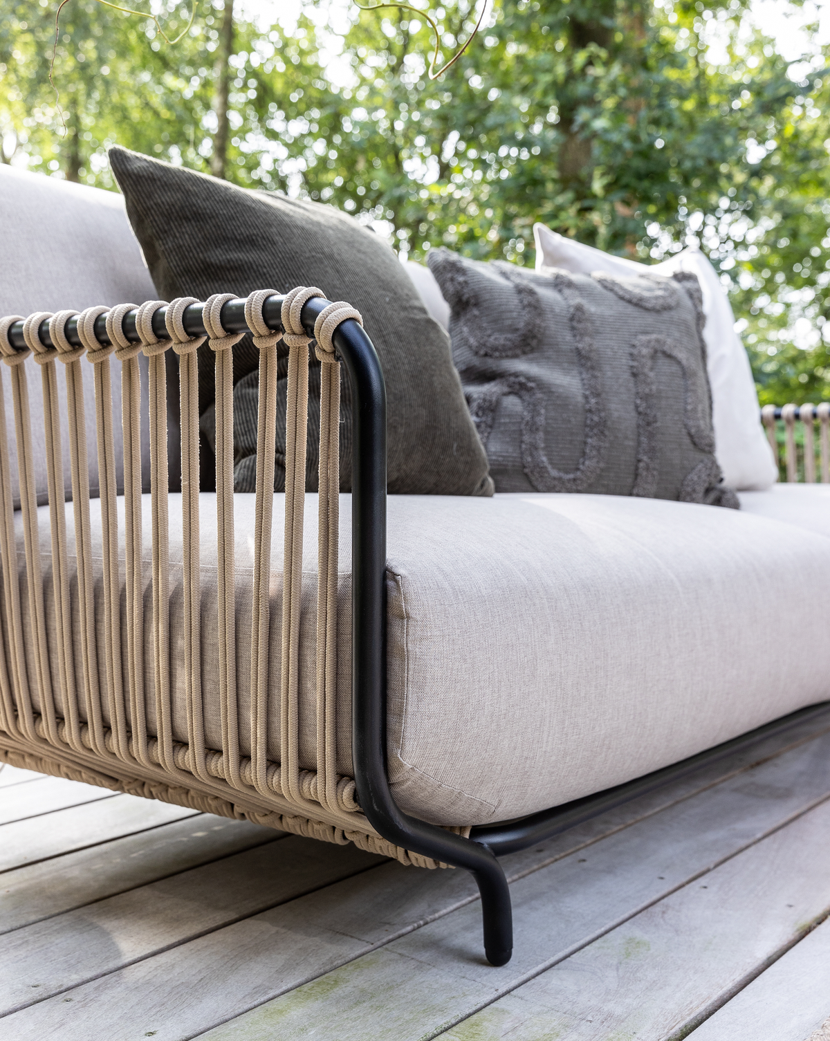 Nærbillede af moderne udendørs sofa med beige snordetaljer og sort metalarm, lyse hynder og mørkegrå puder på en træterrasse i grønne omgivelser.
