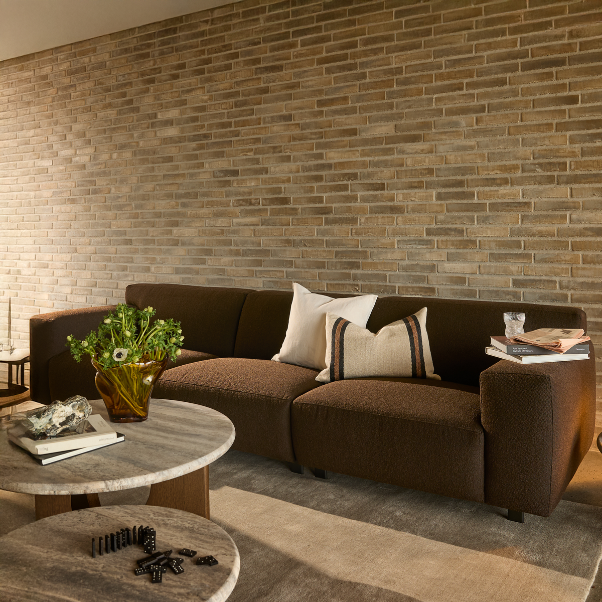 Brun sofa foran lys murstensvæg i stue med to runde stentop-borde, glasvase med grønne blomster, puder, bøger og et glas.