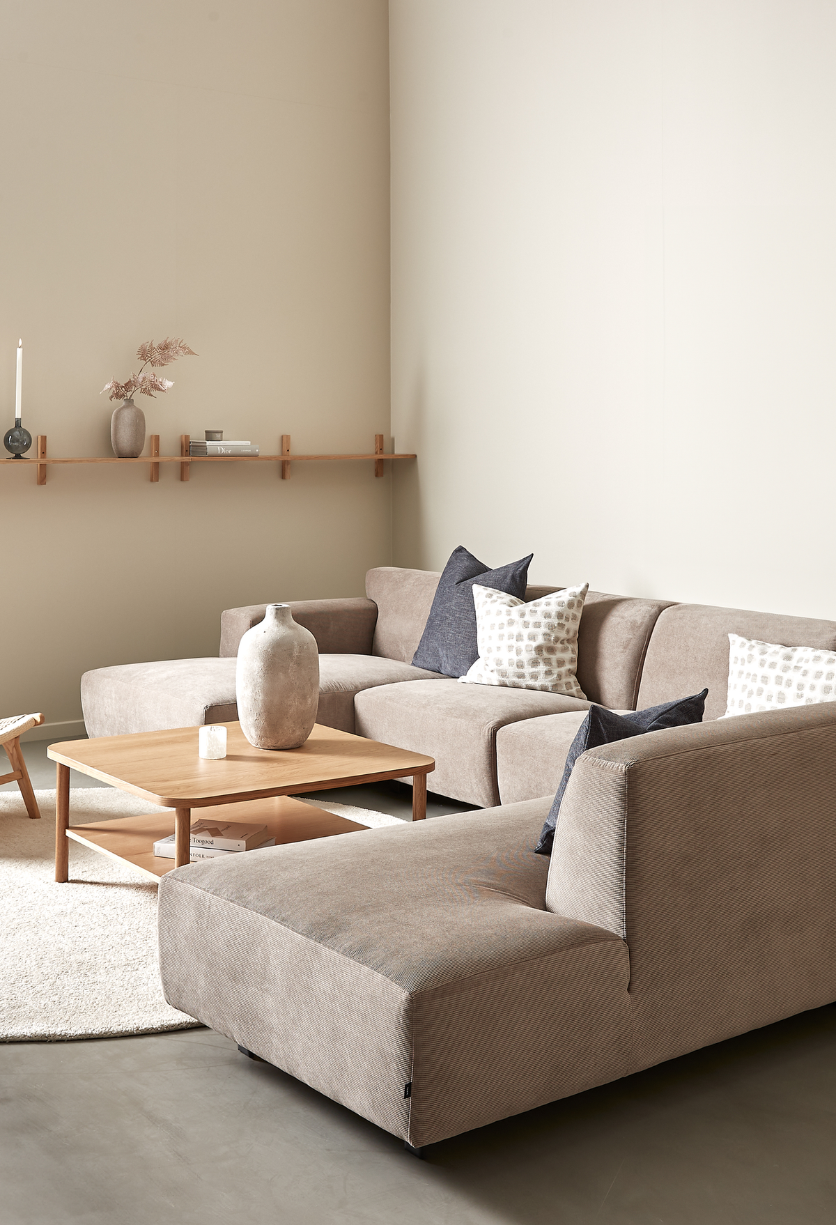 Beige hjørnesofa med træ-sofabord, neutrale puder og væghylde med vase og lys i en lys stue.