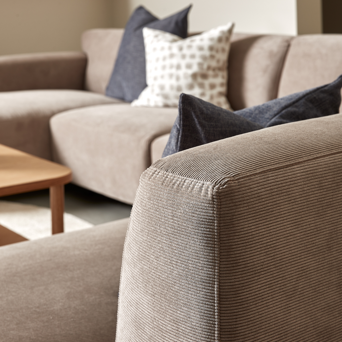 Nærbillede af beige fløjl/cord-sofa med mørkeblå og mønstrede puder ved siden af et lyst træbord i stuen.
