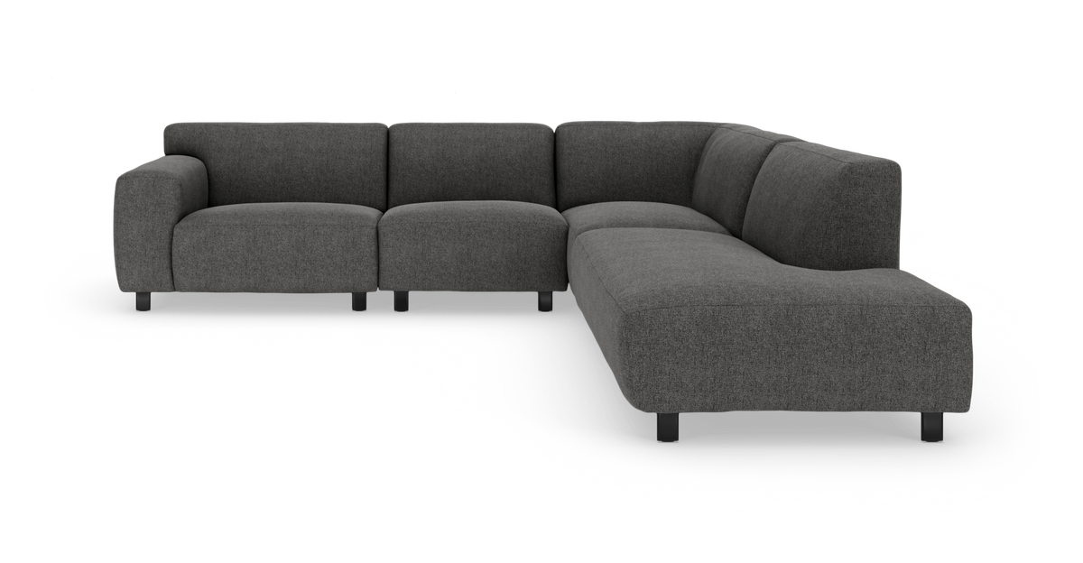 Mørkegrå modulær hjørnesofa i L‑form med afrundede hjørner og lave sorte ben, frilagt på hvid baggrund.