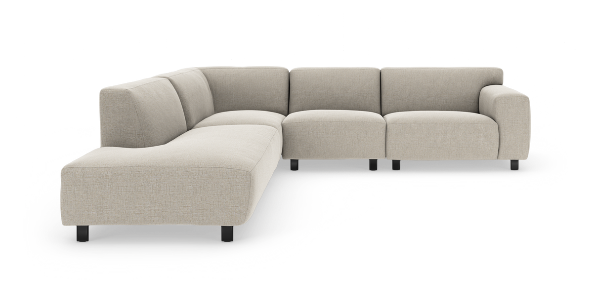 Beige L‑formet modulsofa med bred chaiselong til venstre, bløde afrundede puder og sorte ben, frilagt på hvid baggrund.