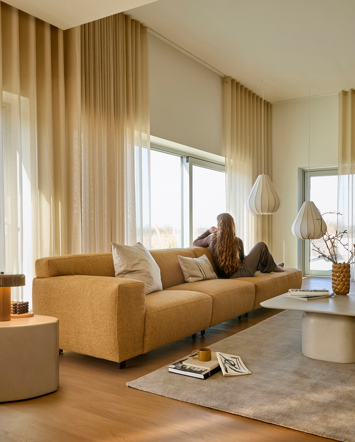 Lyst skandinavisk stue med beige gardiner, sennepsgul sofa med puder, hængende lamper og sofabord med bøger; en person sidder og kigger ud gennem de store vinduer.