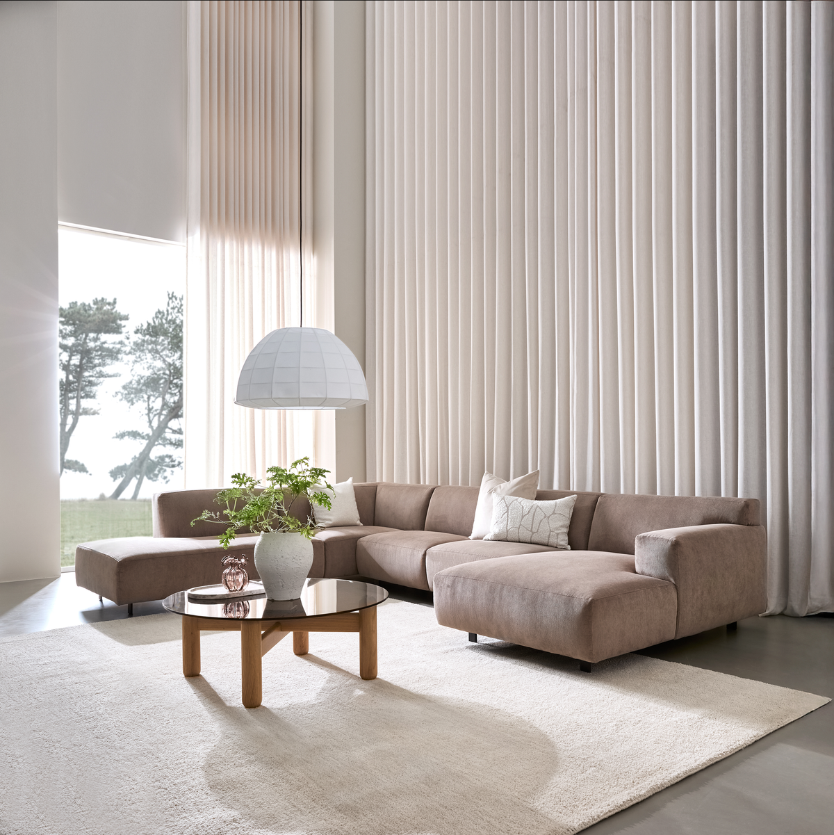 Lys stue med beige hjørnesofa, rundt sofabord i glas med vase og grøn plante, stor hvid pendel og gulvlange lyse gardiner ved et stort vindue.