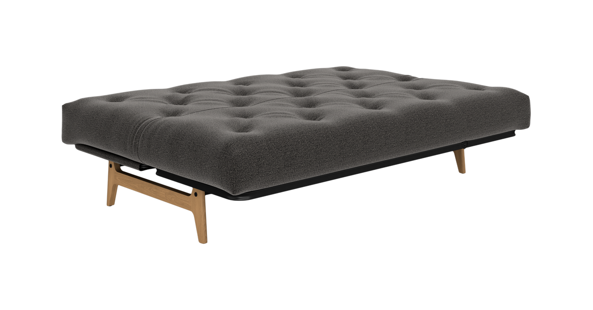 Mørkegrå tuftet sovesofa/futon i tekstil med træben, frilagt på hvid baggrund.