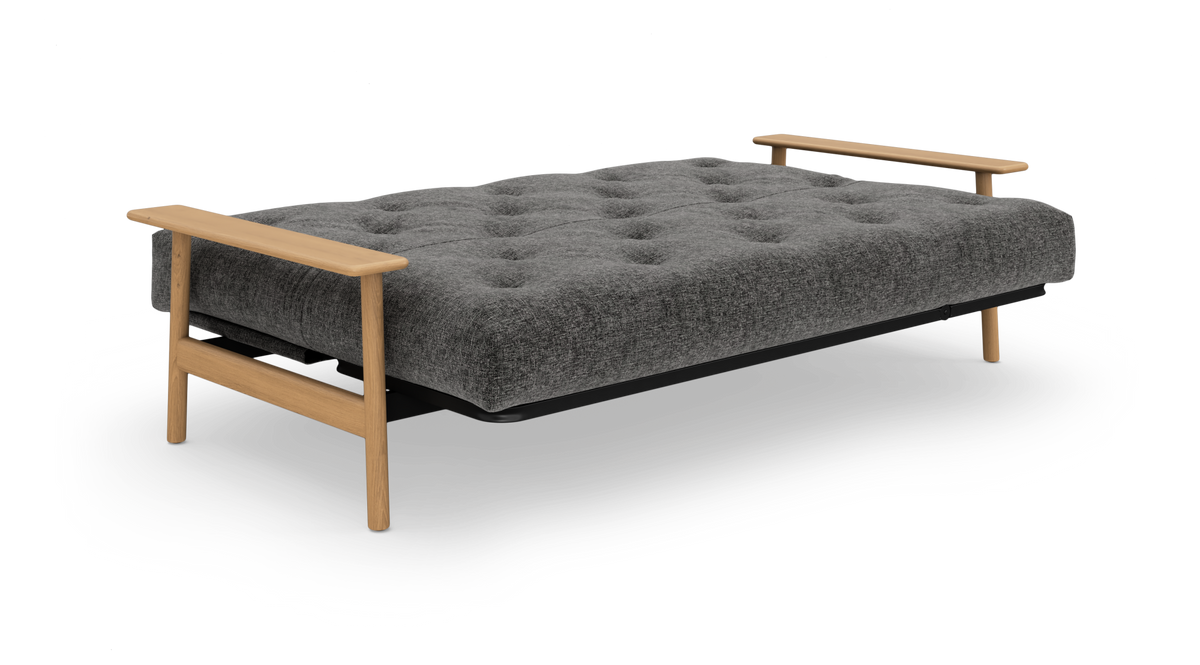 Grå sovesofa med knapstukket madras og lyse træarmlæn, frilagt på hvid baggrund.