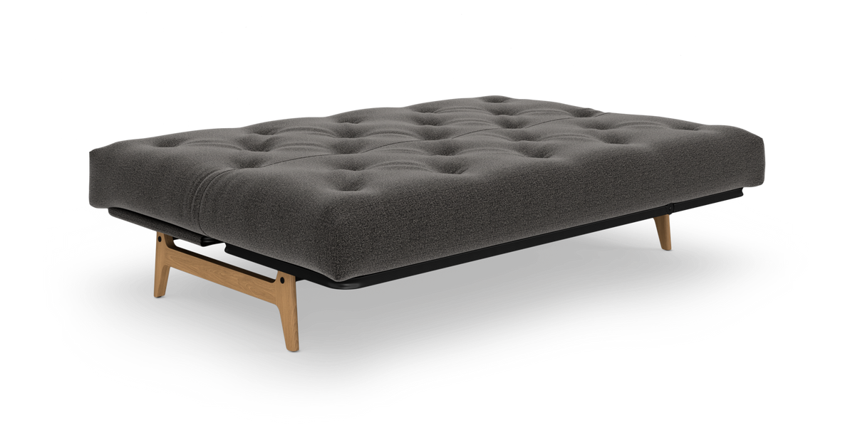 Lav grå tuftet futon/sovesofa med træben, frilagt på hvid baggrund.