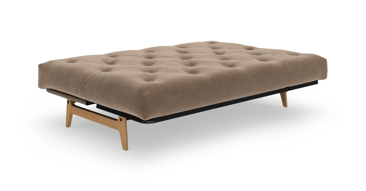 Beige, tuftet sovesofa/futon på træben, frilagt i studie i skrå frontvinkel.